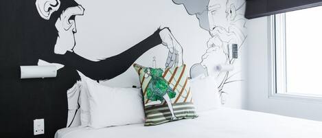 A modern hotel room with a large bed, white bedding, a green and orange patterned pillow, and a black and white mural on the wall.