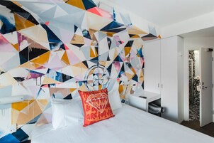 Une chambre d’hôtel moderne avec une fresque murale abstraite aux motifs géométriques, un lit blanc avec un oreiller rouge et une petite table de chevet.