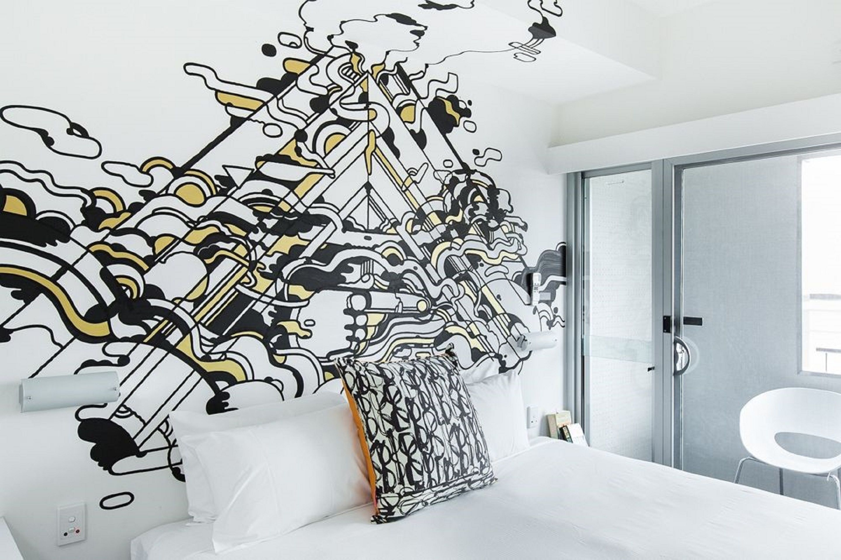 Una habitació d'hotel moderna amb un mural abstracte gran a la paret, un llit blanc i una porta de vidre que dóna a un balcó.