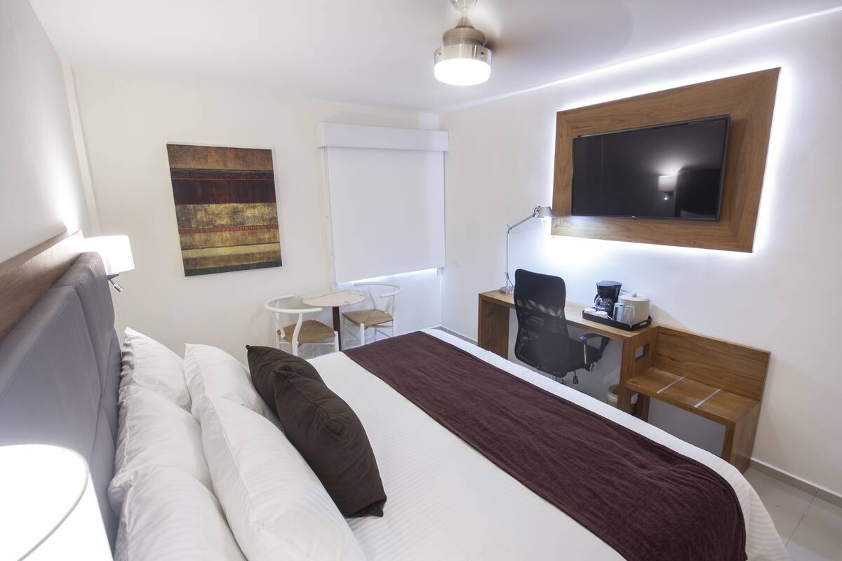A hotel room with a bed, a desk, a chair, a TV, and a painting on the wall.