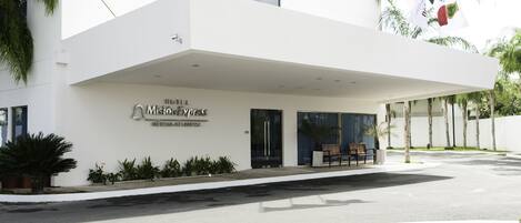 A modern hotel entrance with a covered walkway, palm trees, and a sign indicating it is the "Hotel Mision de Jesus."