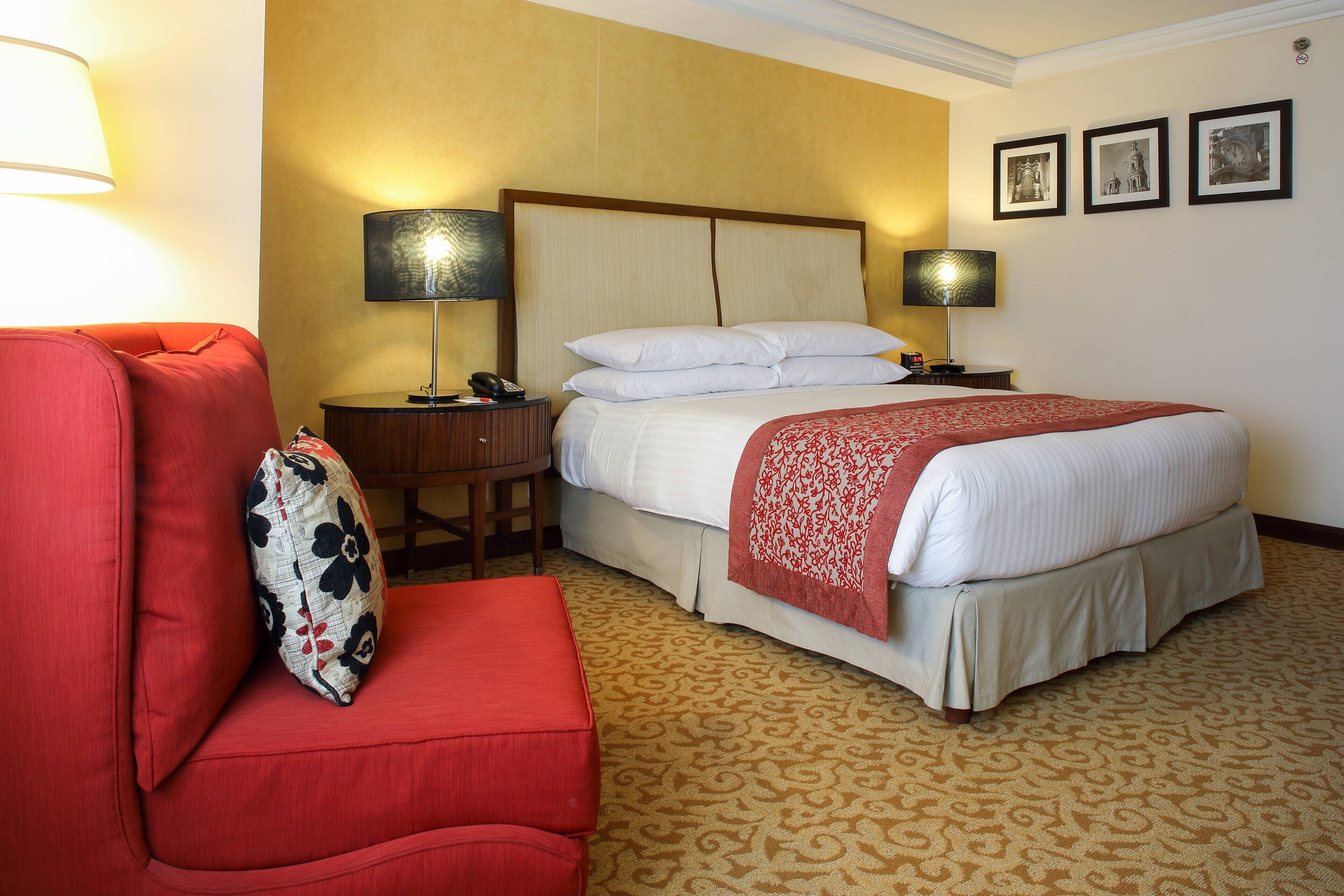 A hotel room with a bed, a red armchair, a nightstand, and a lamp.