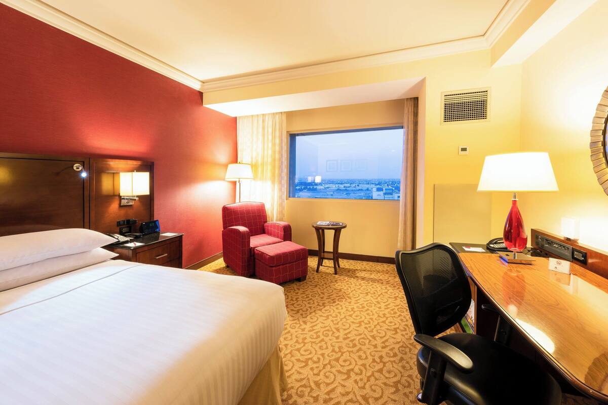 A hotel room with a bed, a desk, a chair, a small table, and a window with a city view.