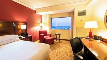 A hotel room with a bed, a desk, a chair, a small table, and a window with a city view.
