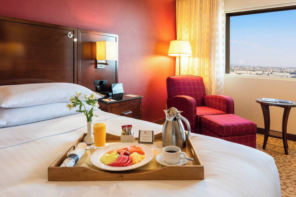 A hotel room with a bed, a tray with breakfast, a coffee cup, a lamp, and a view of the city.