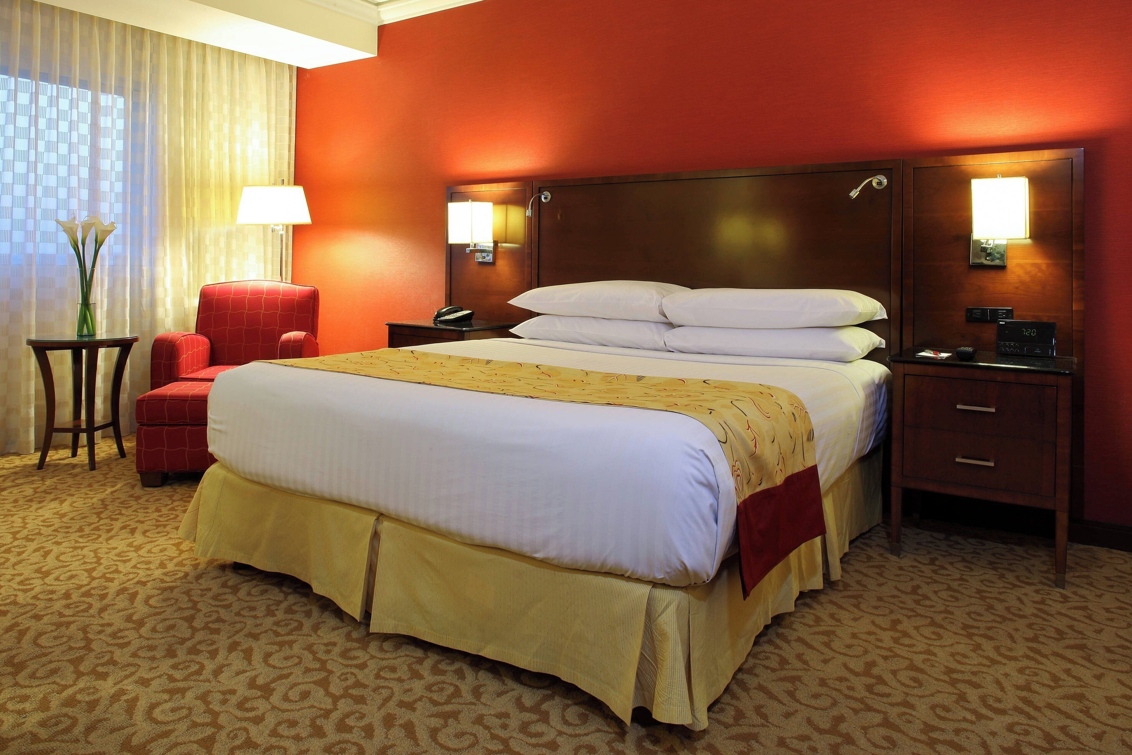 A hotel room with a large bed, a red armchair, a nightstand with a lamp, and a bedside table with a clock.