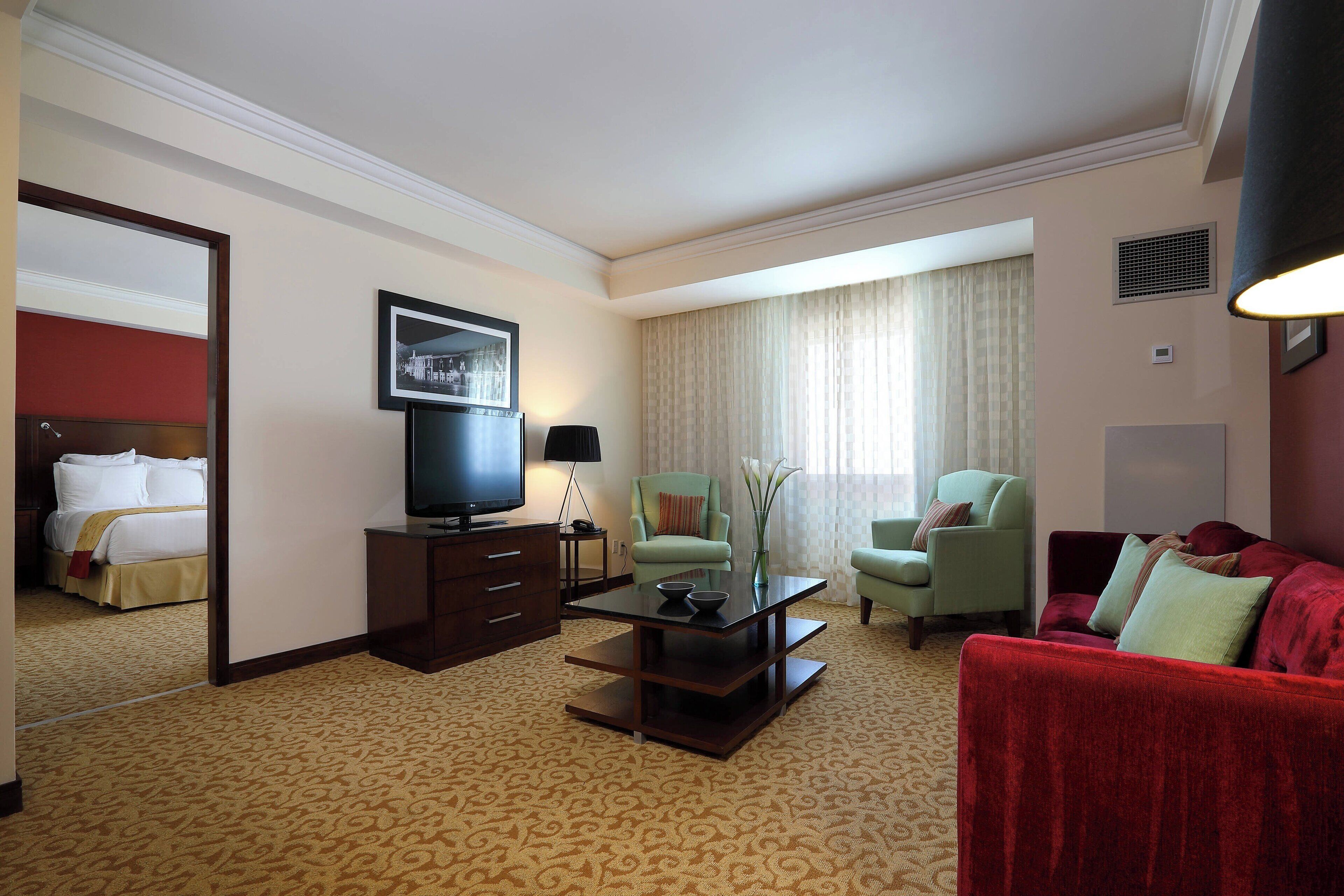 Habitación de hotel con cama, televisor, mesa de centro, dos sillones, un sofá rojo y un amplio ventanal con cortinas.