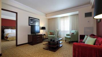 Habitación de hotel con cama, televisor, mesa de centro, dos sillones, un sofá rojo y un amplio ventanal con cortinas.