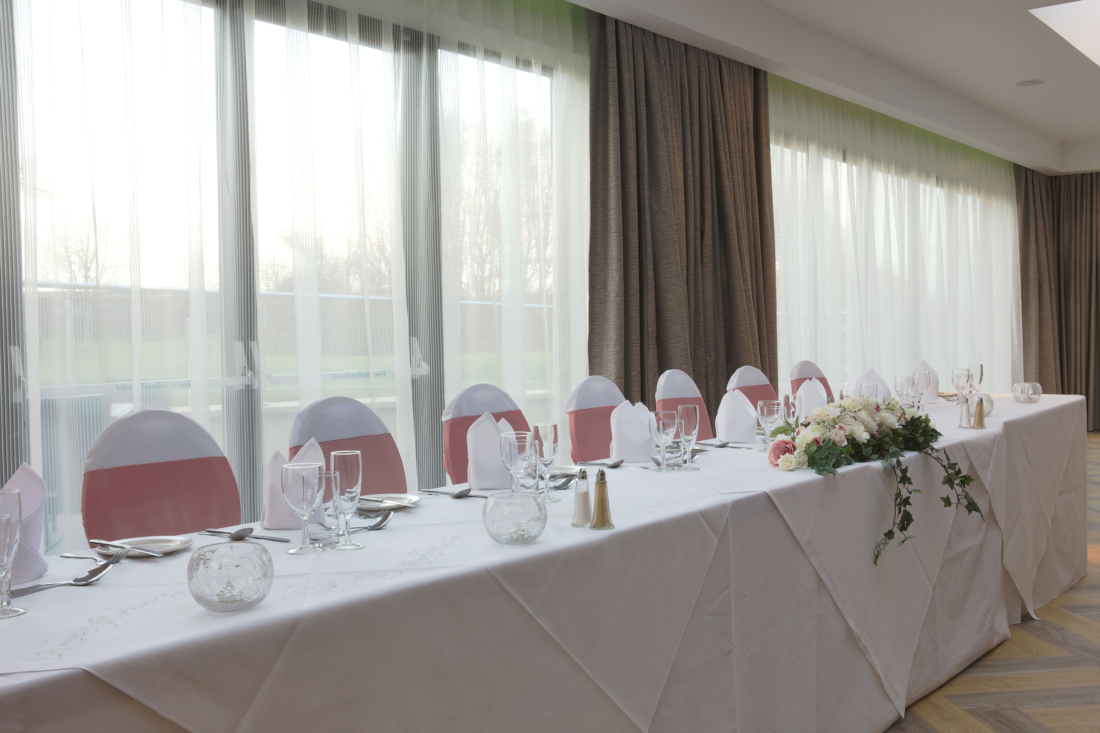 A banquet table set for a formal event with white tablecloths, pink and white chairs, and floral centerpieces.