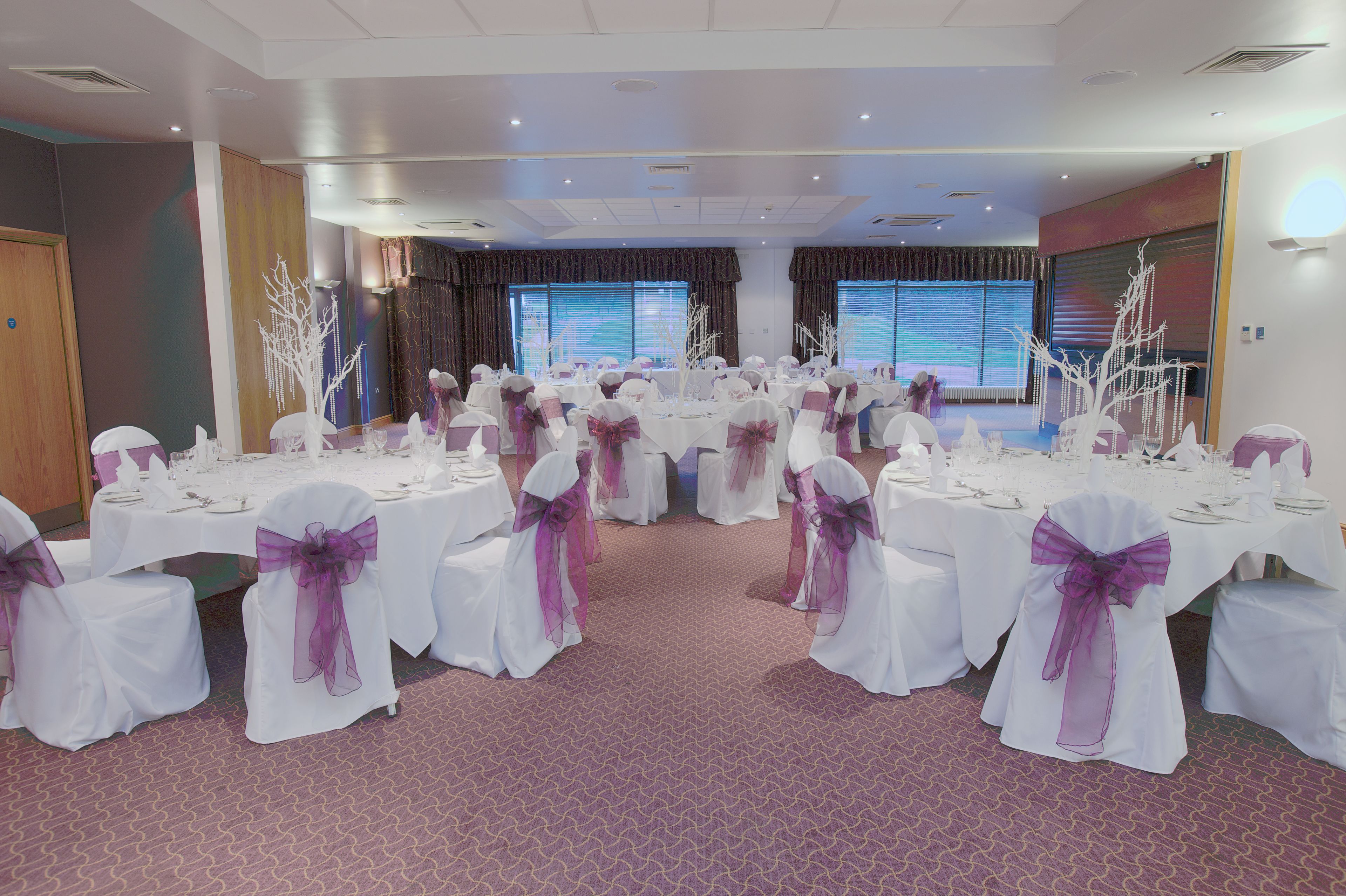 Un salón de banquetes con mesas redondas cubiertas con manteles blancos y sillas adornadas con cintas moradas, dispuestas en filas.