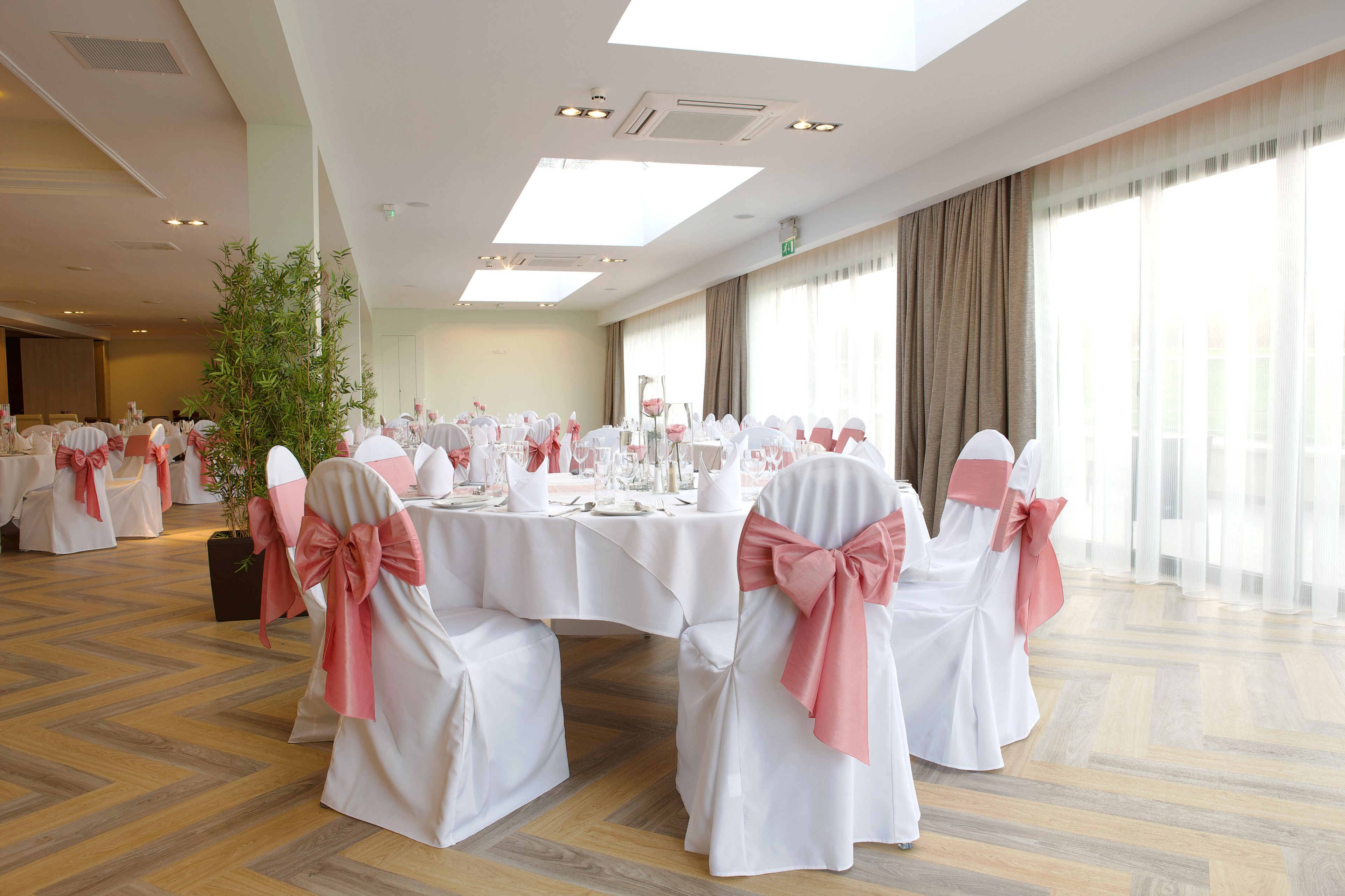 A banquet hall with round tables set for a formal event, white chairs with pink sashes, and large windows with sheer curtains.