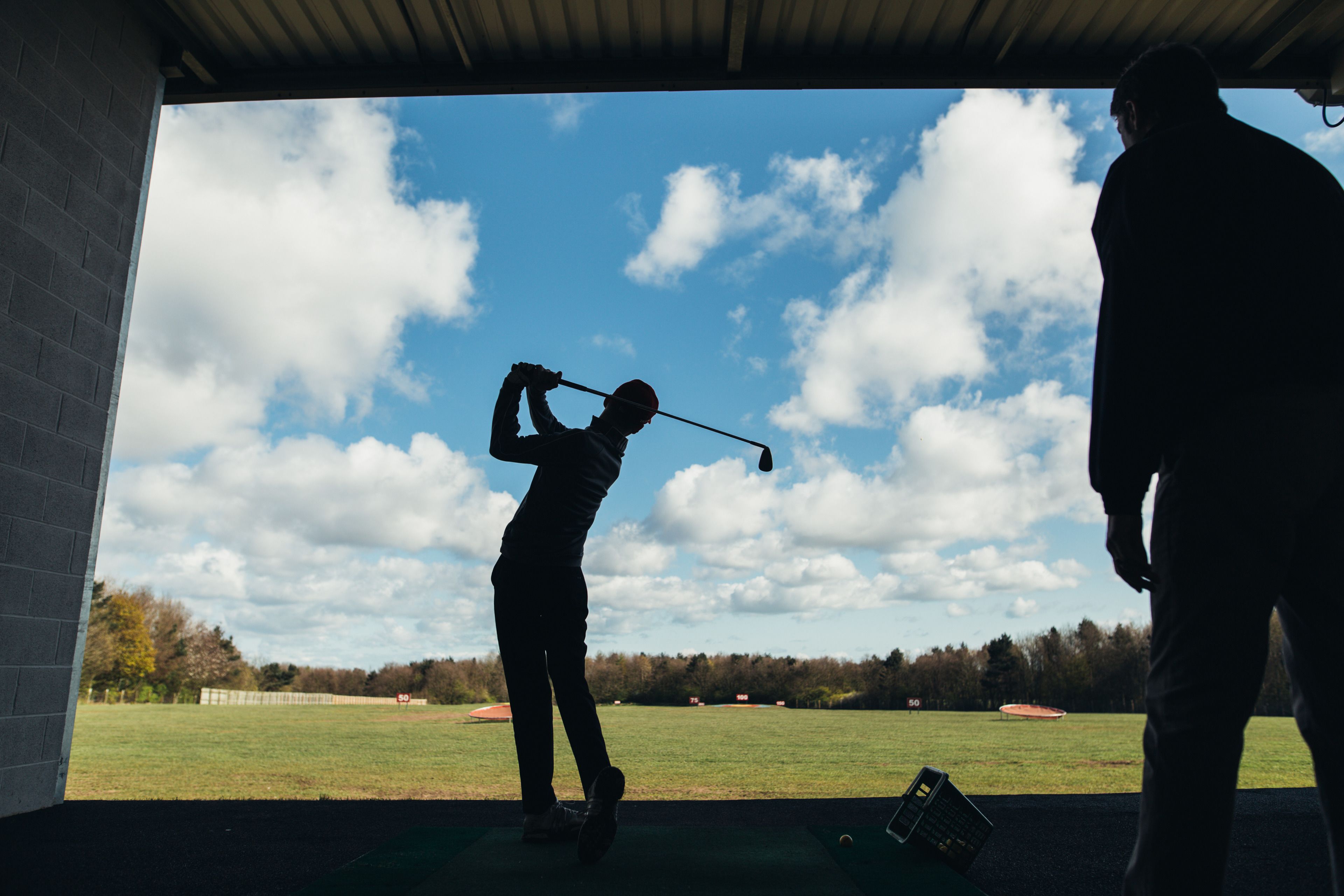 Silueta de una persona jugando golf dentro de un área techada, con cielo despejado y árboles al fondo.