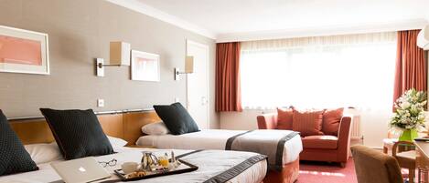 A hotel room with two beds, a sofa, a desk with a chair, a laptop on one bed, and a vase with flowers on the desk.