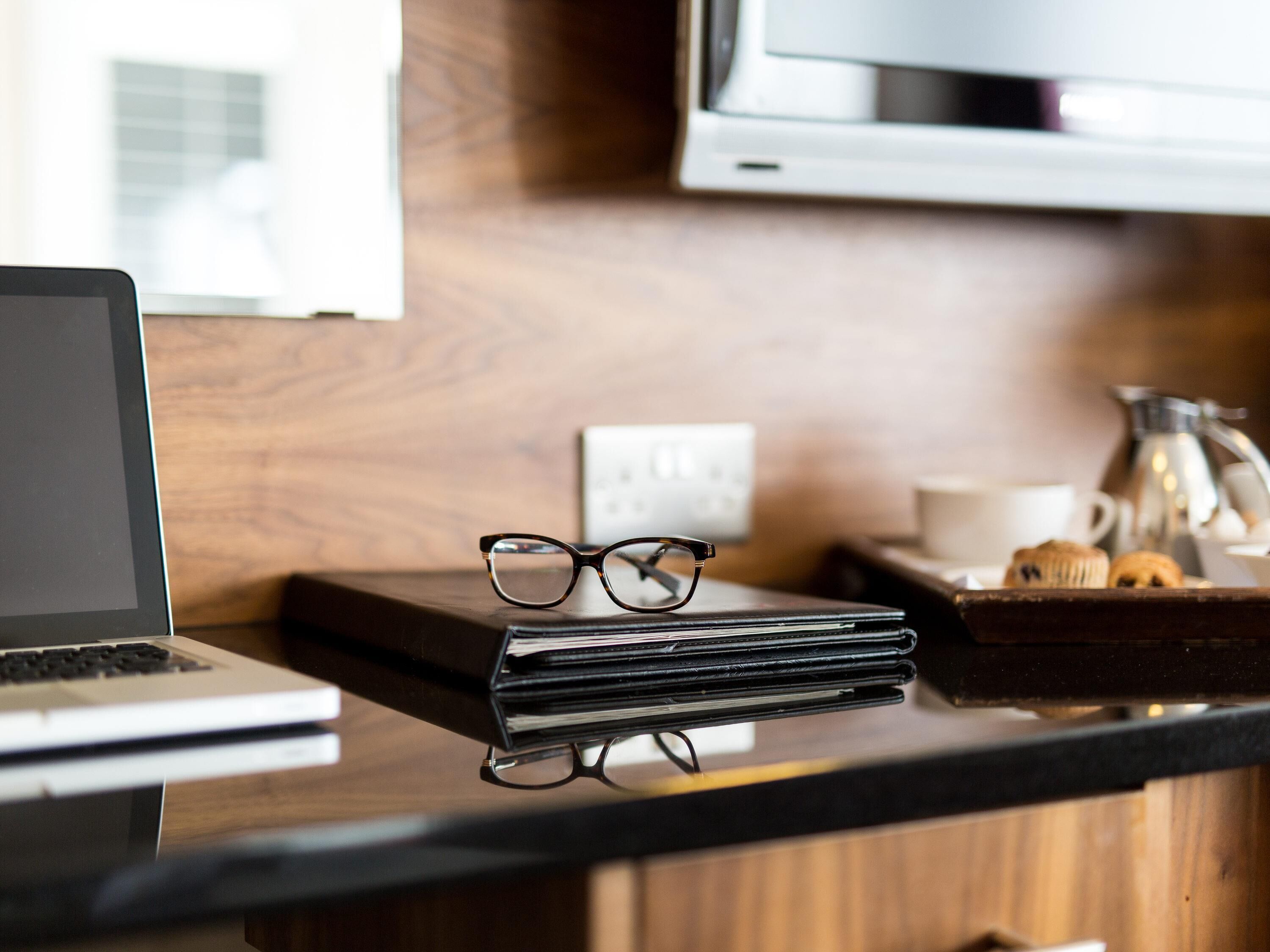 A laptop, a pair of glasses, a stack of books, a coffee maker, and a muffin on a kitchen counter.