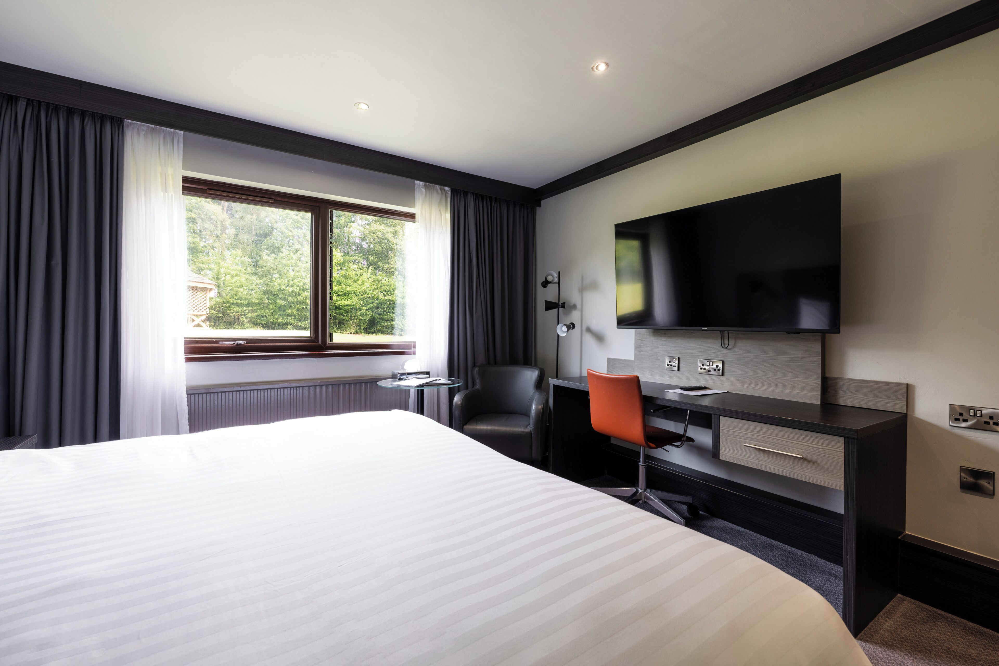 A hotel room with a large bed, a desk with a chair, a flat-screen TV mounted on the wall, and a window with curtains.