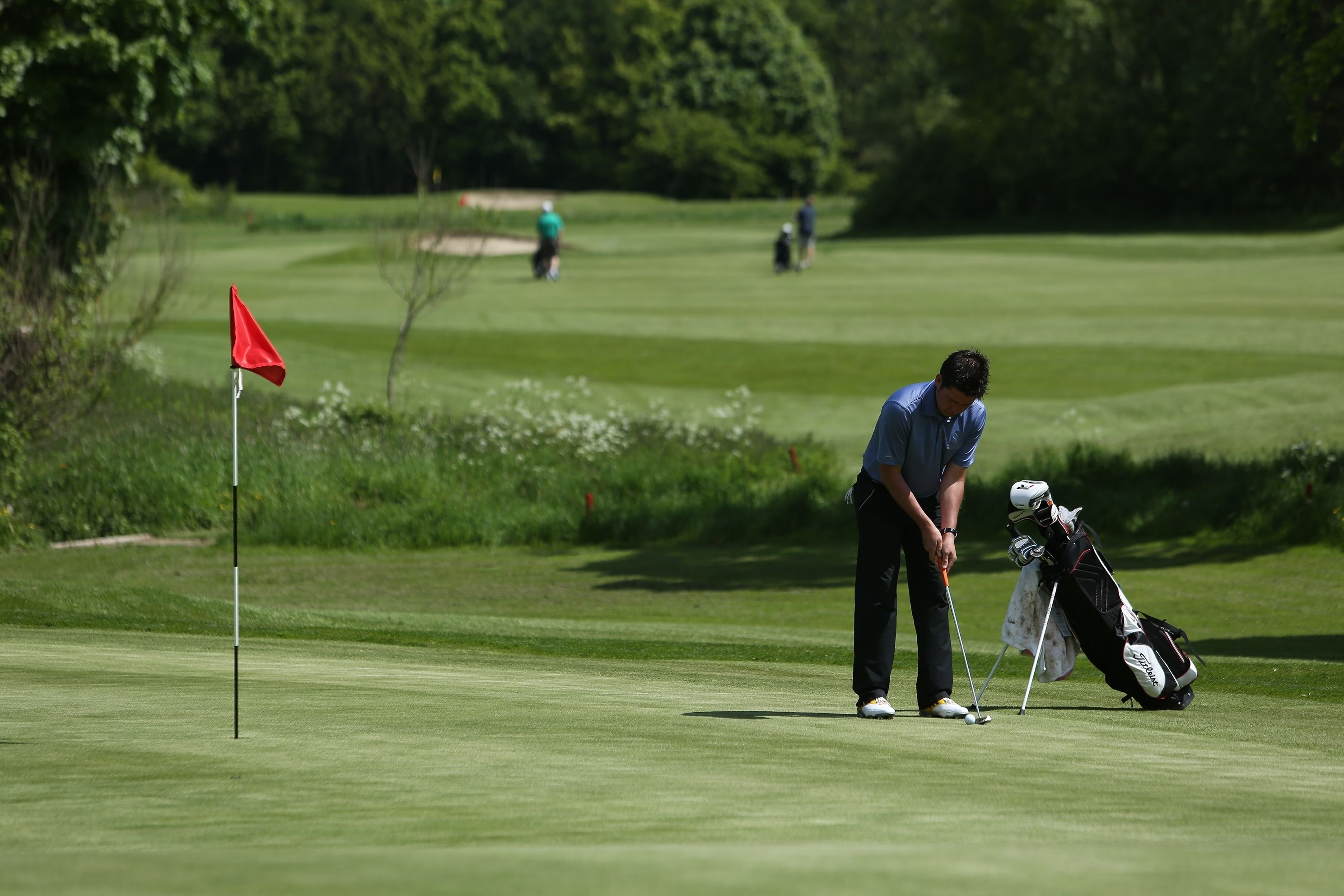 A golf course with a red flag, a golfer preparing to putt, and a golf bag.