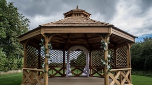 Un belvédère en bois, surmonté d’un toit de tuiles, entouré de verdure et sous un ciel nuageux.