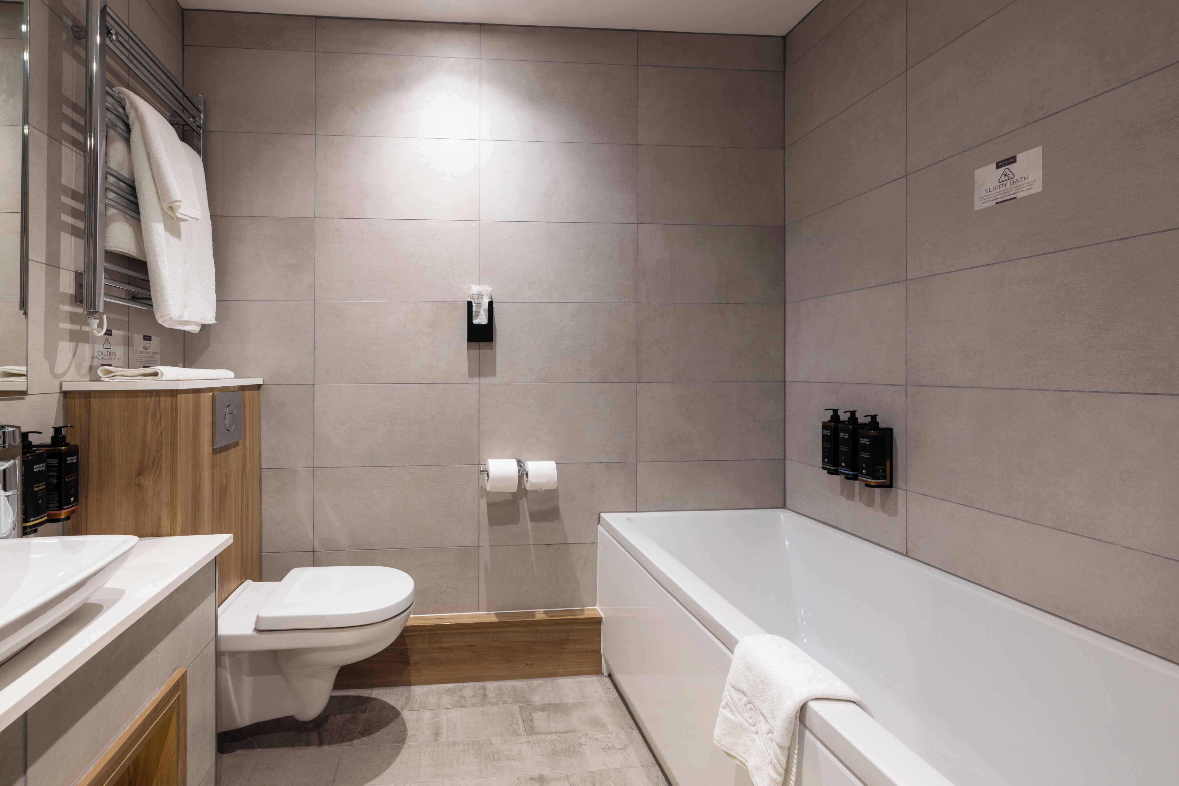 A modern bathroom with a toilet, sink, and bathtub. There are towels hanging on a rack, and toiletries are placed on the wall.