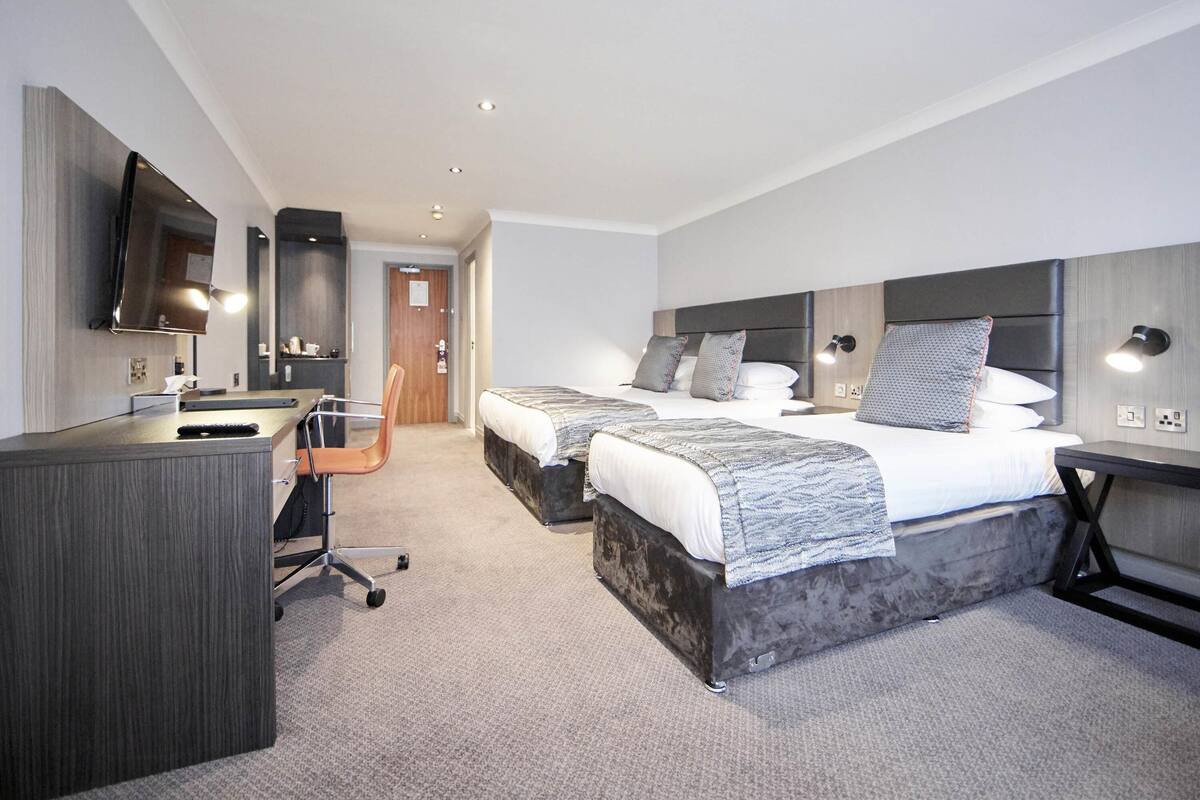 A modern hotel room with a desk, two beds, a TV, and a bathroom.