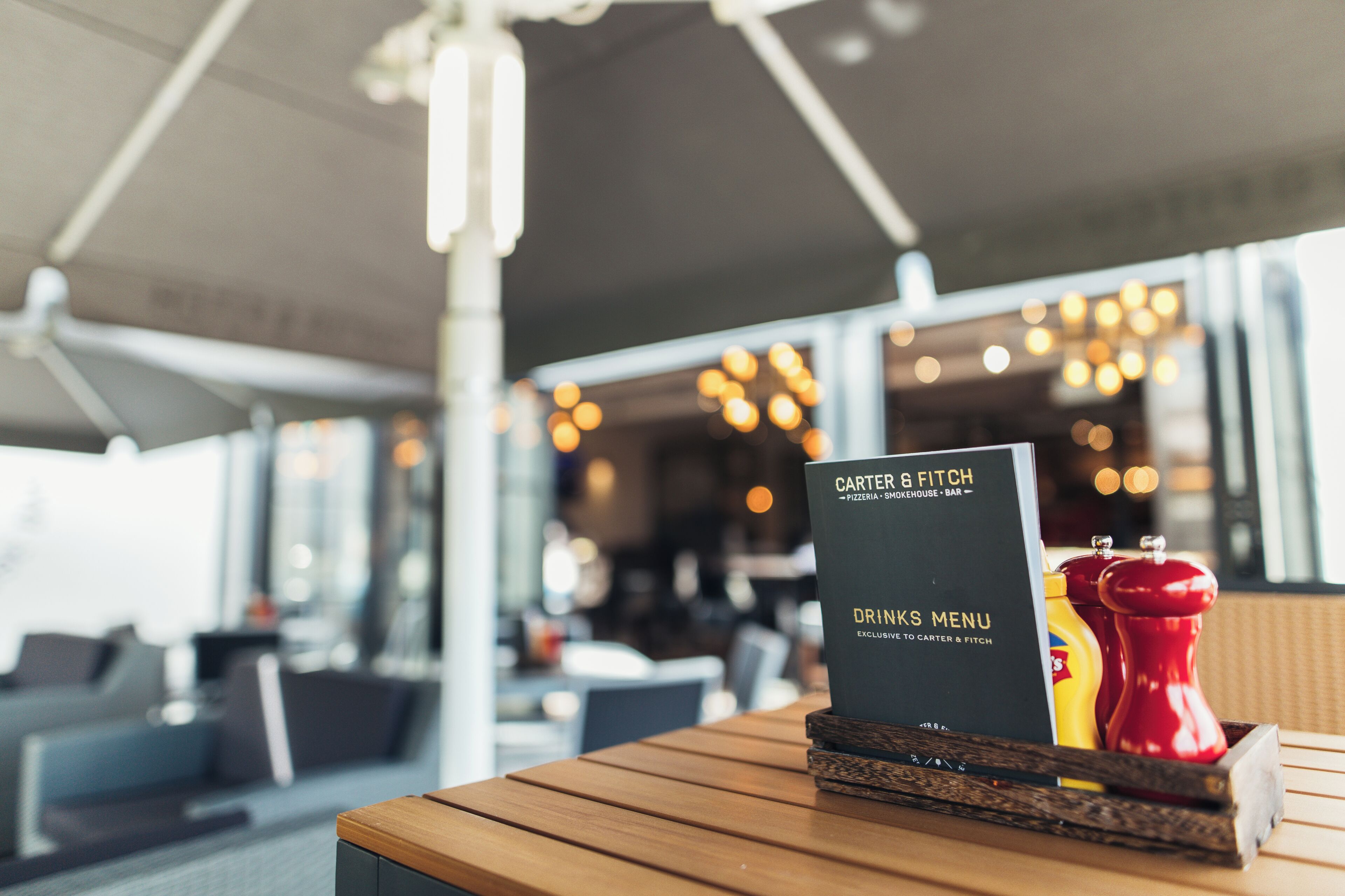 Un atril con un menú de bebidas, una botella de kétchup y un molinillo de pimiento rojo sobre una mesa de madera en un restaurante.