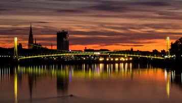 Vista nocturna de un puente con luces iluminadas que se reflejan en el agua, una torre destacada y un edificio moderno al fondo.