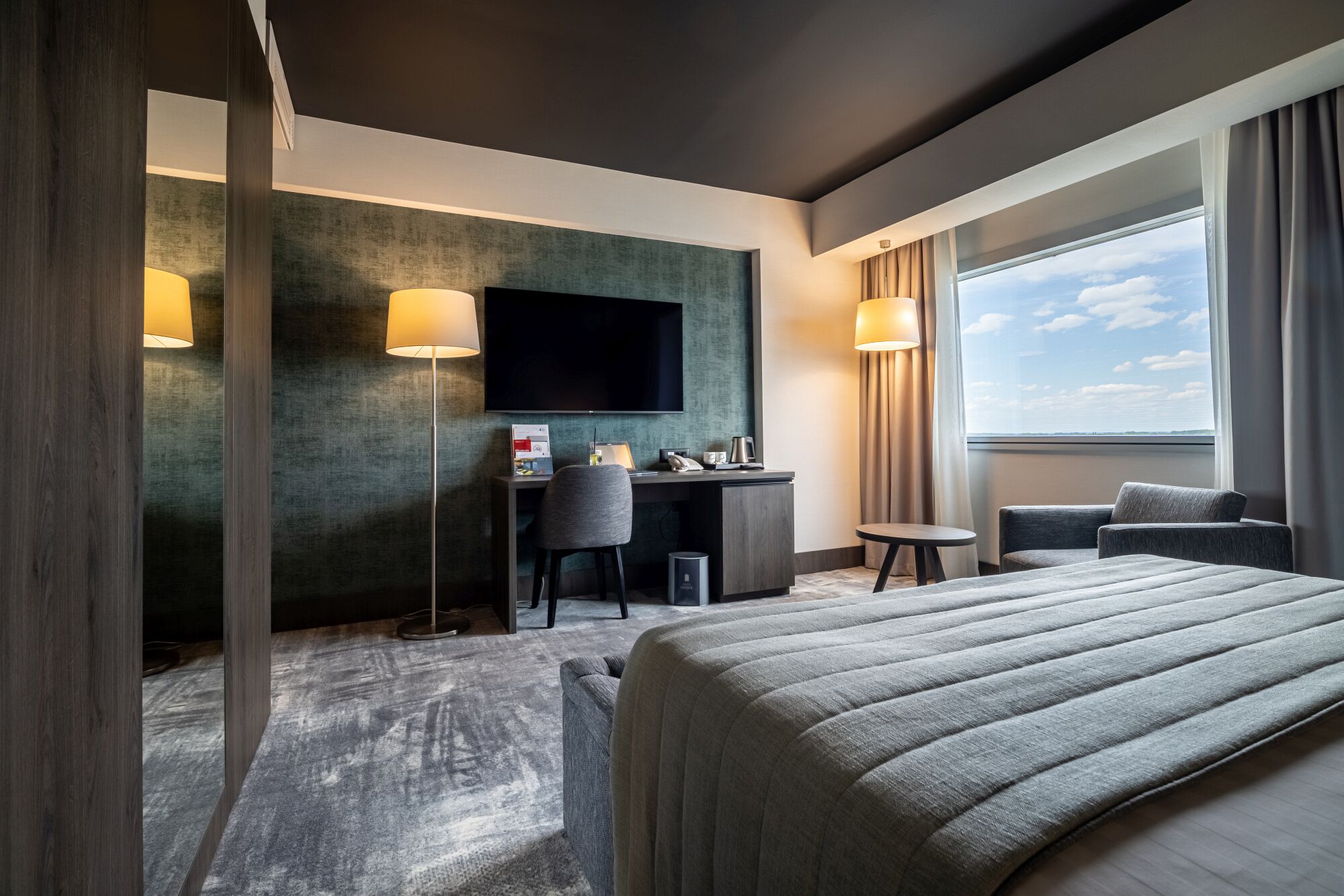 A hotel room with a bed, a desk with a chair, a TV, and a window with curtains.