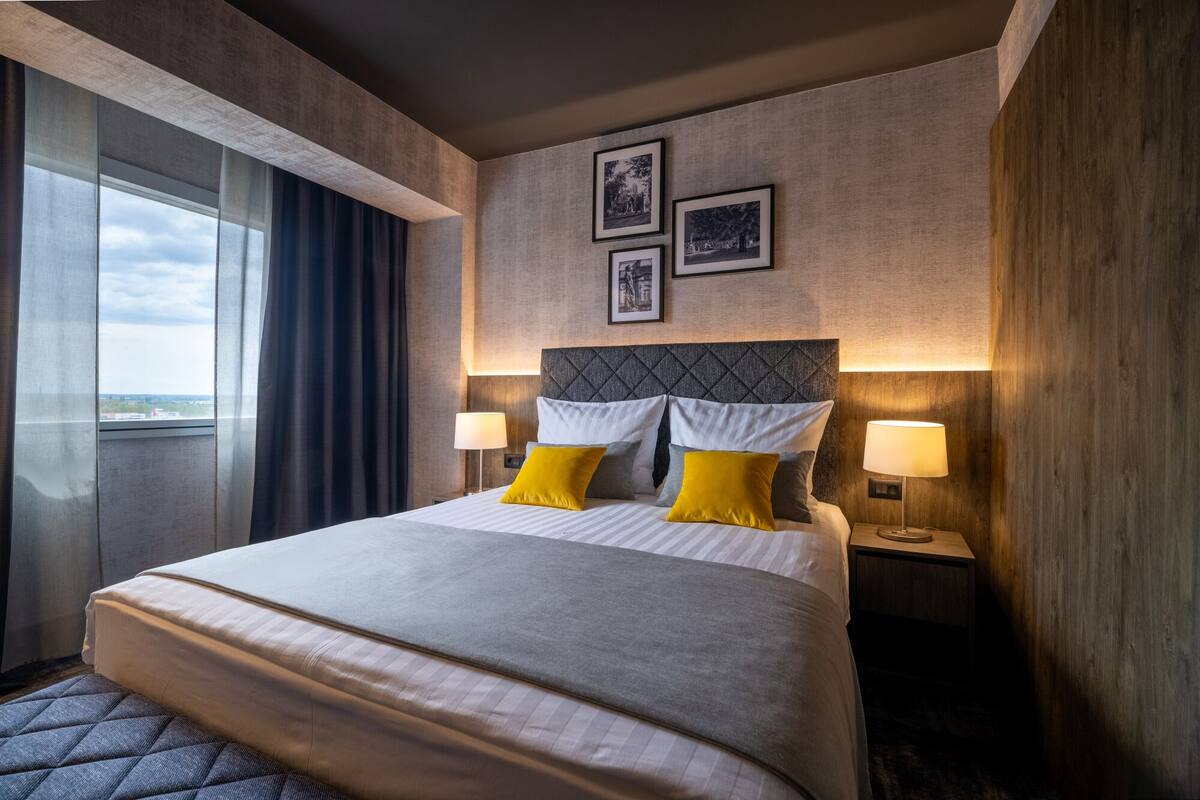 Habitación de hotel con una cama grande, dos lámparas de noche, vistas a través de una ventana y dos cuadros en la pared.