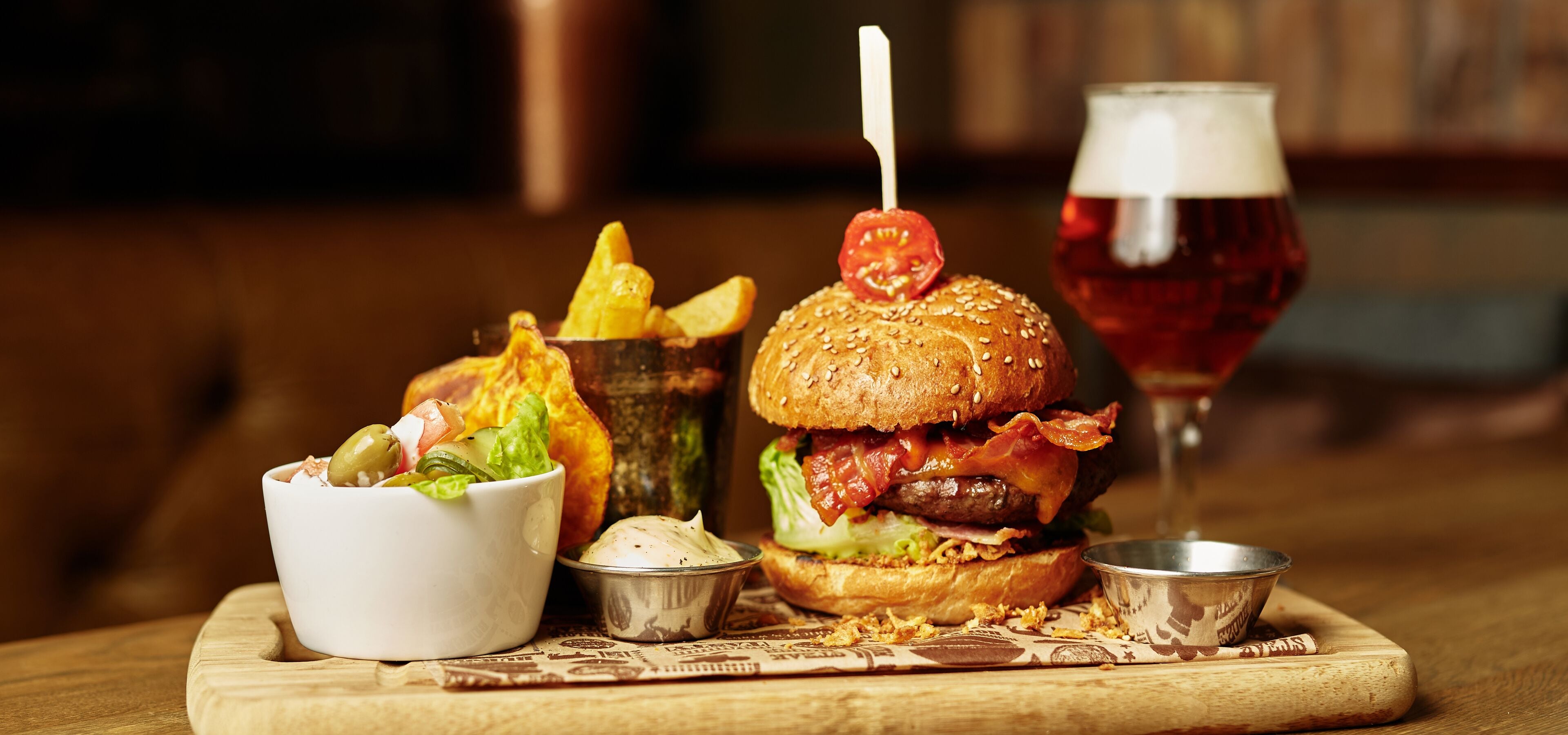Una hamburguesa con tocino, lechuga y tomate en un pan, acompañada de papas fritas, una ensalada y una cerveza en una mesa de madera.