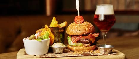 Una hamburguesa con tocino, lechuga y tomate en un pan, acompañada de papas fritas, una ensalada y una cerveza en una mesa de madera.