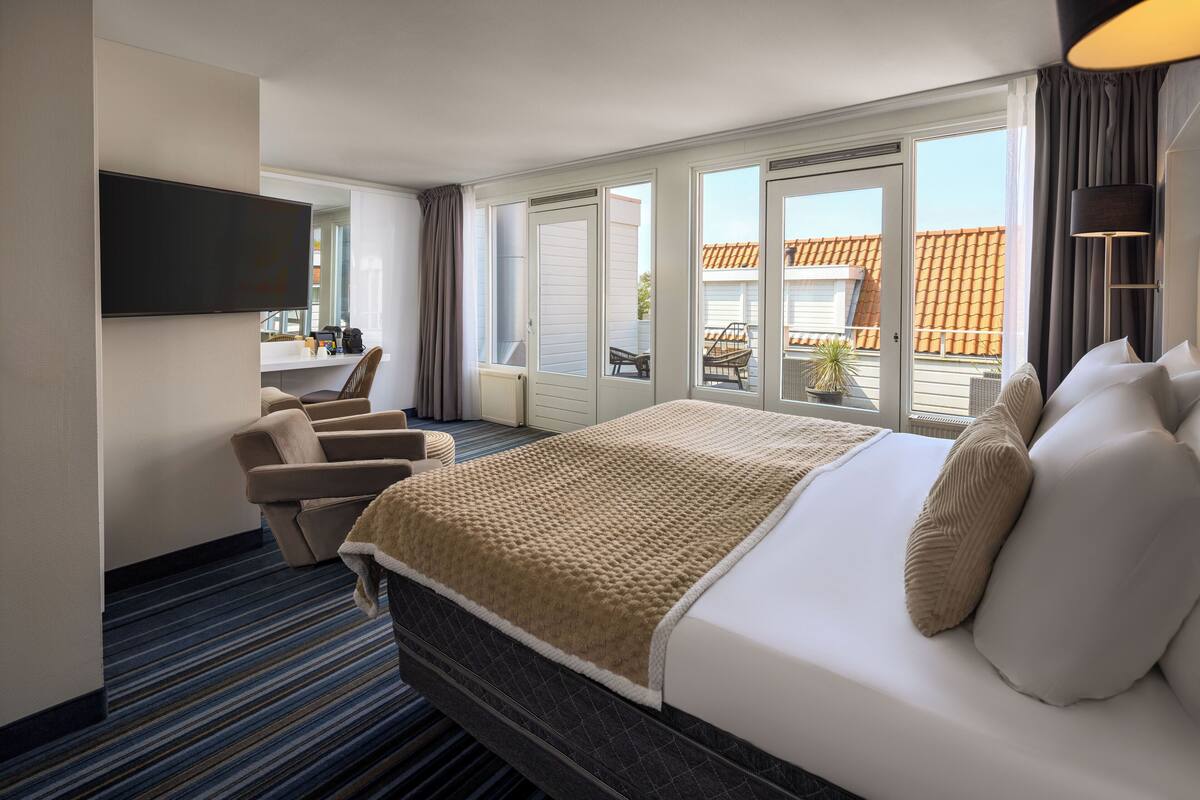 Habitación de hotel con una cama grande, un televisor montado en la pared, una zona de estar con sillas y vistas al exterior a través de las ventanas.