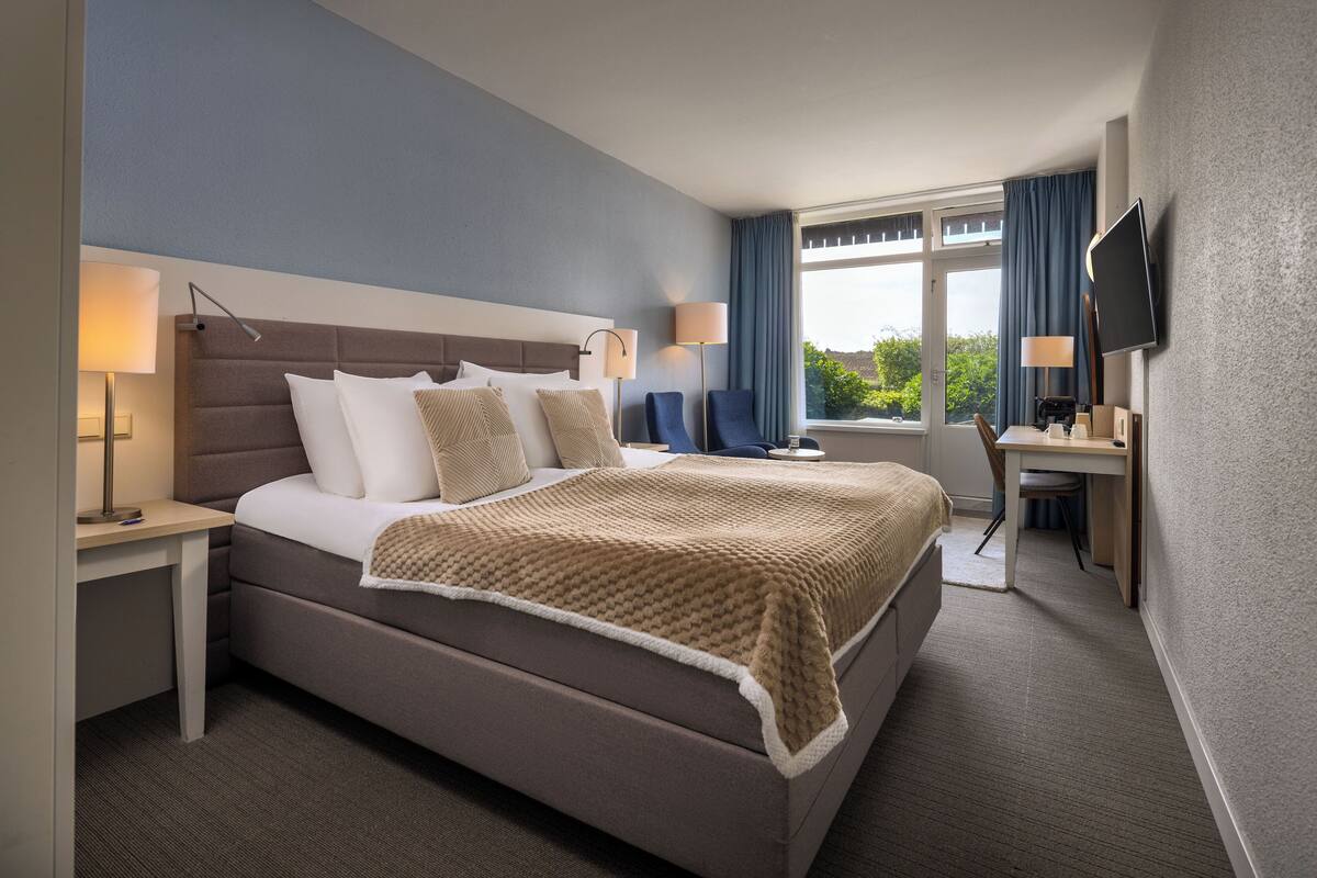 Habitación de hotel con una cama grande, un escritorio, una silla, un televisor y una ventana con cortinas.