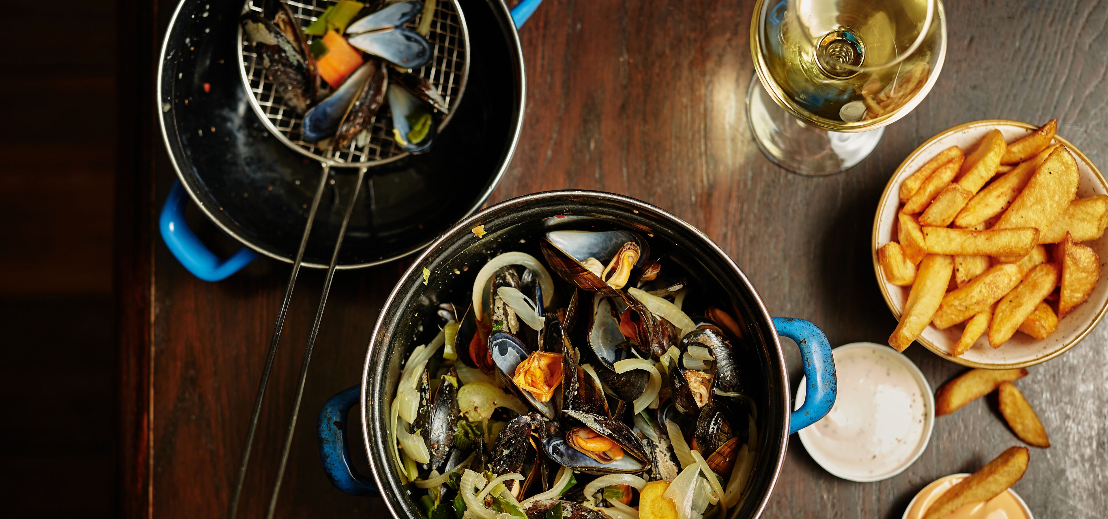 Un plato de mariscos con mejillones, cebollas y limón, acompañado de papas fritas y una copa de vino blanco sobre una mesa de madera.