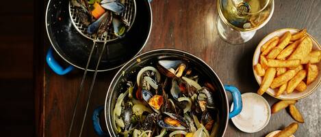 Un plato de mariscos con mejillones, cebollas y limón, acompañado de papas fritas y una copa de vino blanco sobre una mesa de madera.