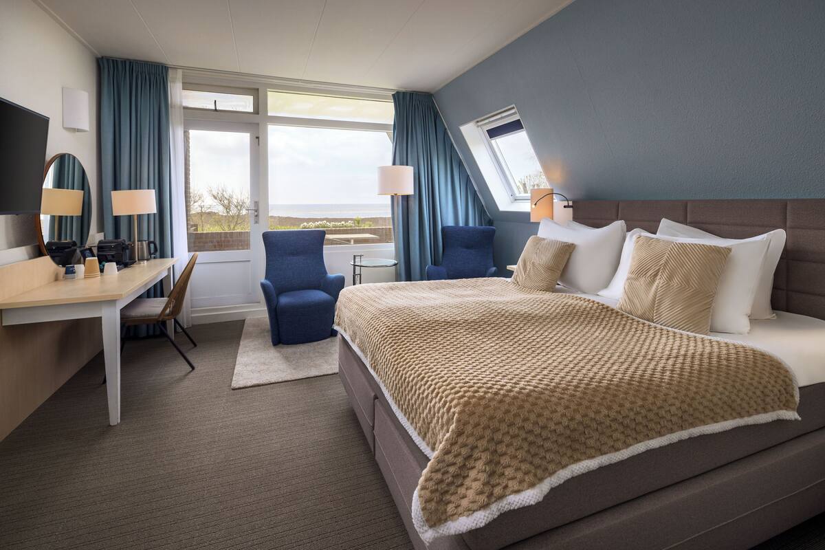 Habitación de hotel con una cama grande, un escritorio con silla, una computadora y vistas al exterior.