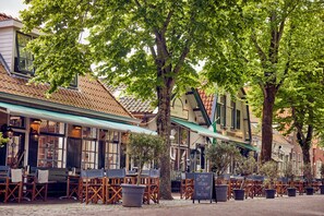 Un café en plein air avec des tables et des chaises en bois, entouré d’arbres et de bâtiments à l’architecture traditionnelle.