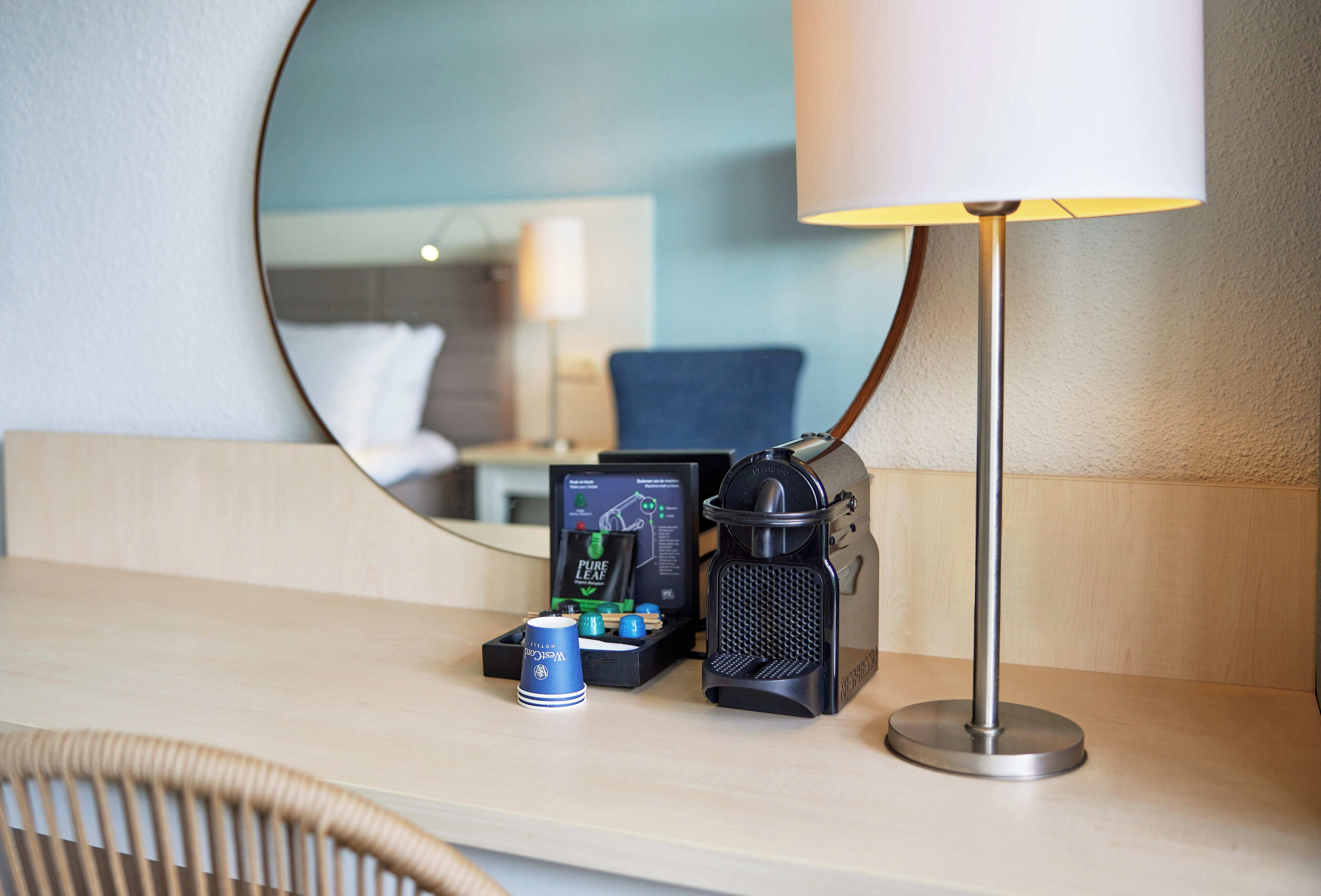 Quarto de hotel com um espelho redondo, uma luminária, uma cafeteira e uma pequena mesa com uma xícara e uma caixa de lenços.