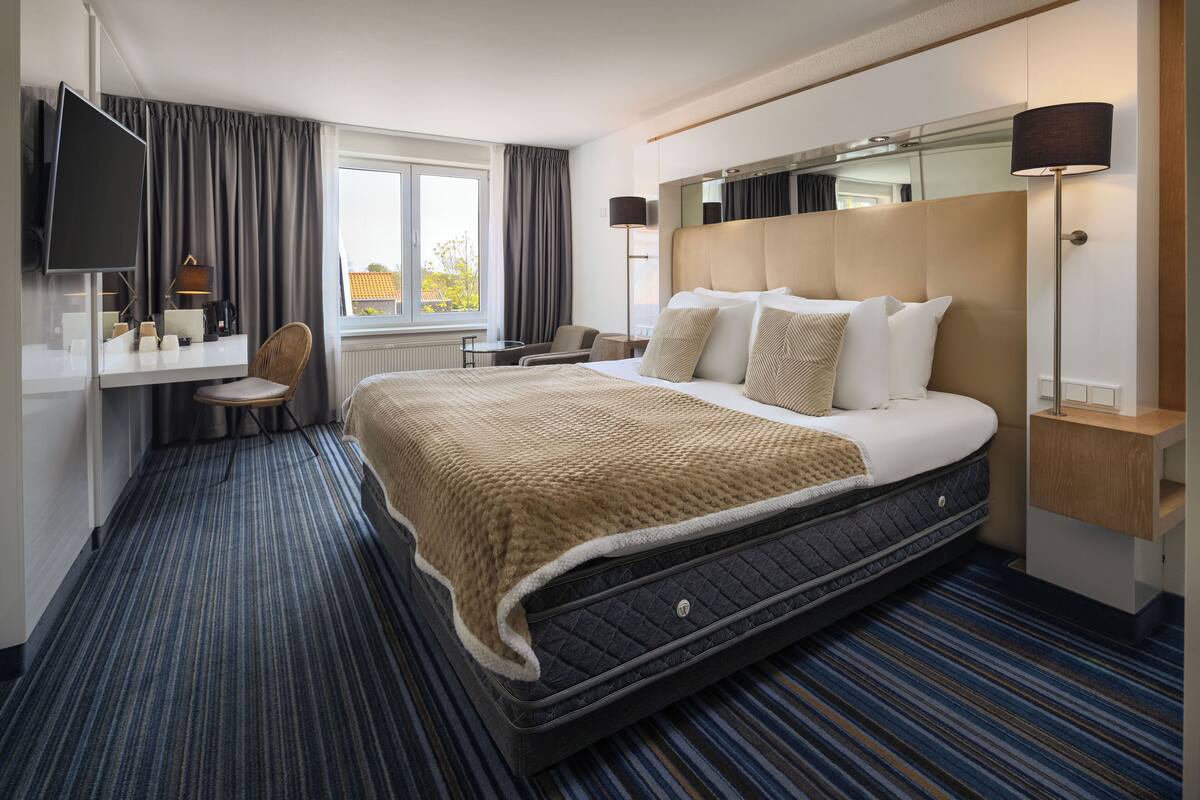 Habitación de hotel con una cama grande, un escritorio con silla, televisión y ventana con cortinas.