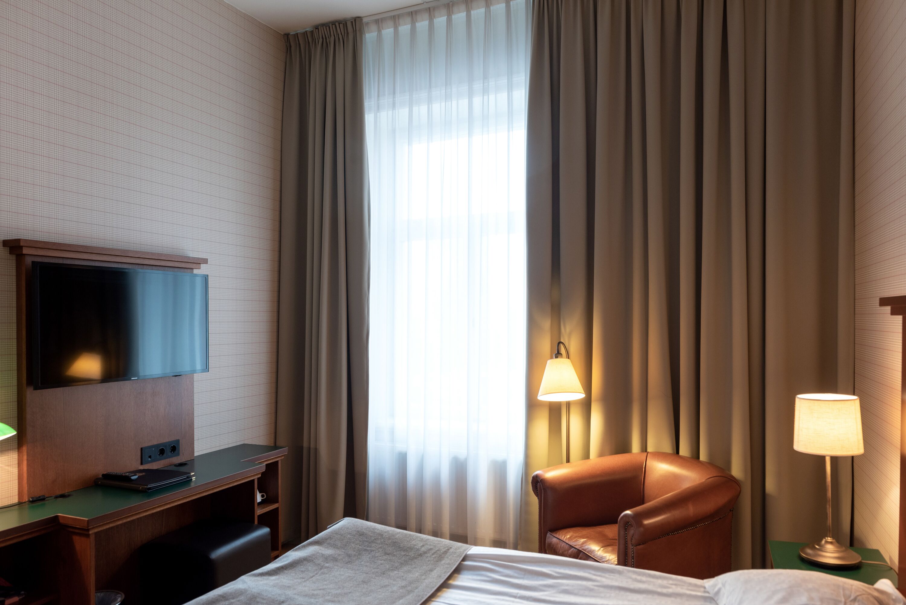 Ett hotellrum med en säng, ett skrivbord med en platt-TV, en fåtölj i läder och en golvlampa.