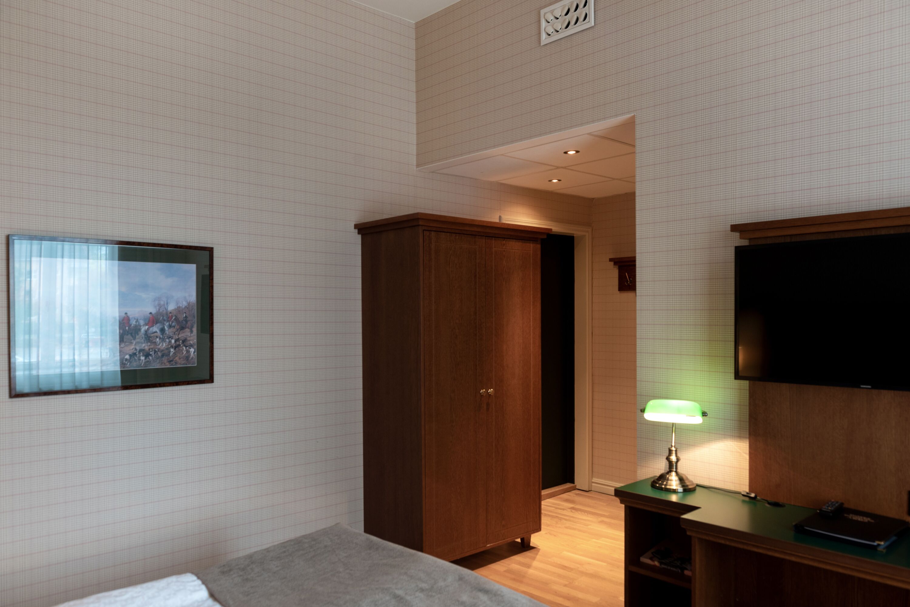 Quarto de hotel com uma cama, guarda-roupa, escrivaninha iluminada por uma luminária e televisão na parede.