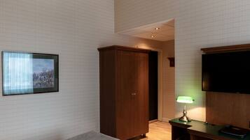 Quarto de hotel com uma cama, guarda-roupa, escrivaninha iluminada por uma luminária e televisão na parede.