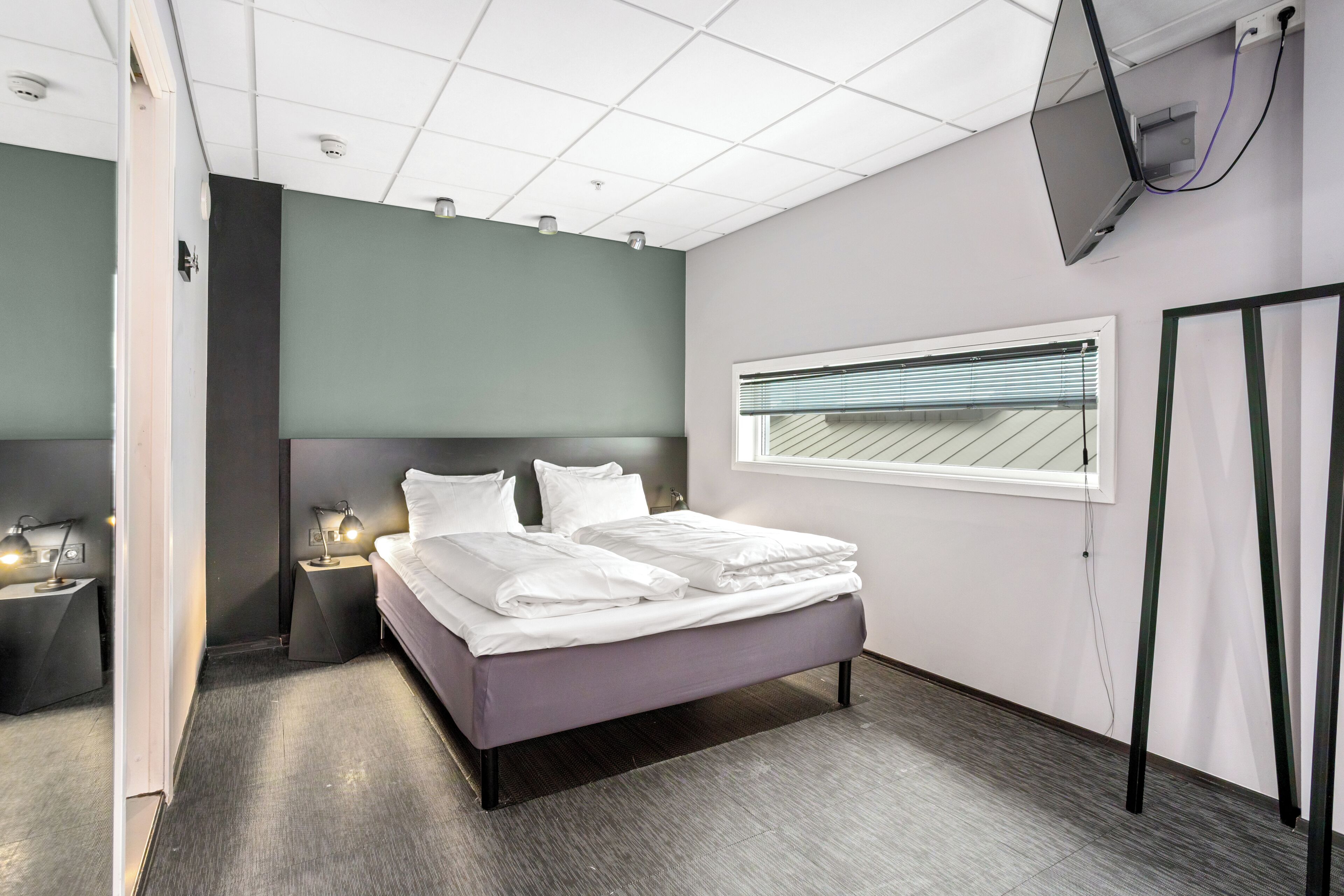 A hotel room with a bed, bedside tables, a TV, a window with blinds, and a door.