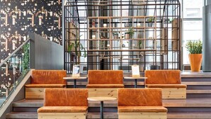 A modern indoor seating area with wooden steps and orange cushioned chairs, a metal grid structure with plants, and a cityscape view through large windows. - Comfort Hotel Union Brygge - Drammen (Drammen)