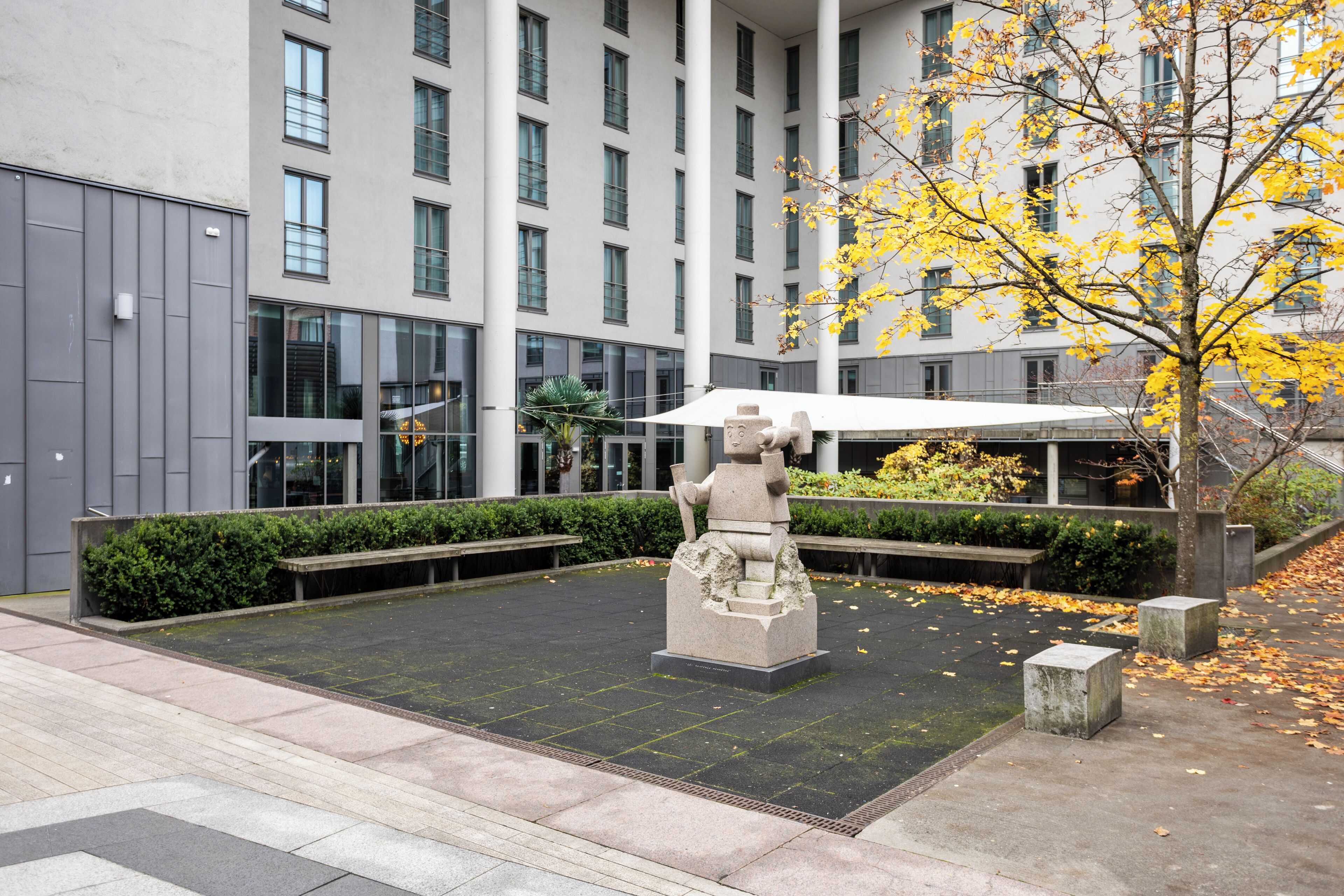A courtyard with a stone sculpture, benches, and a building with multiple windows in the background.