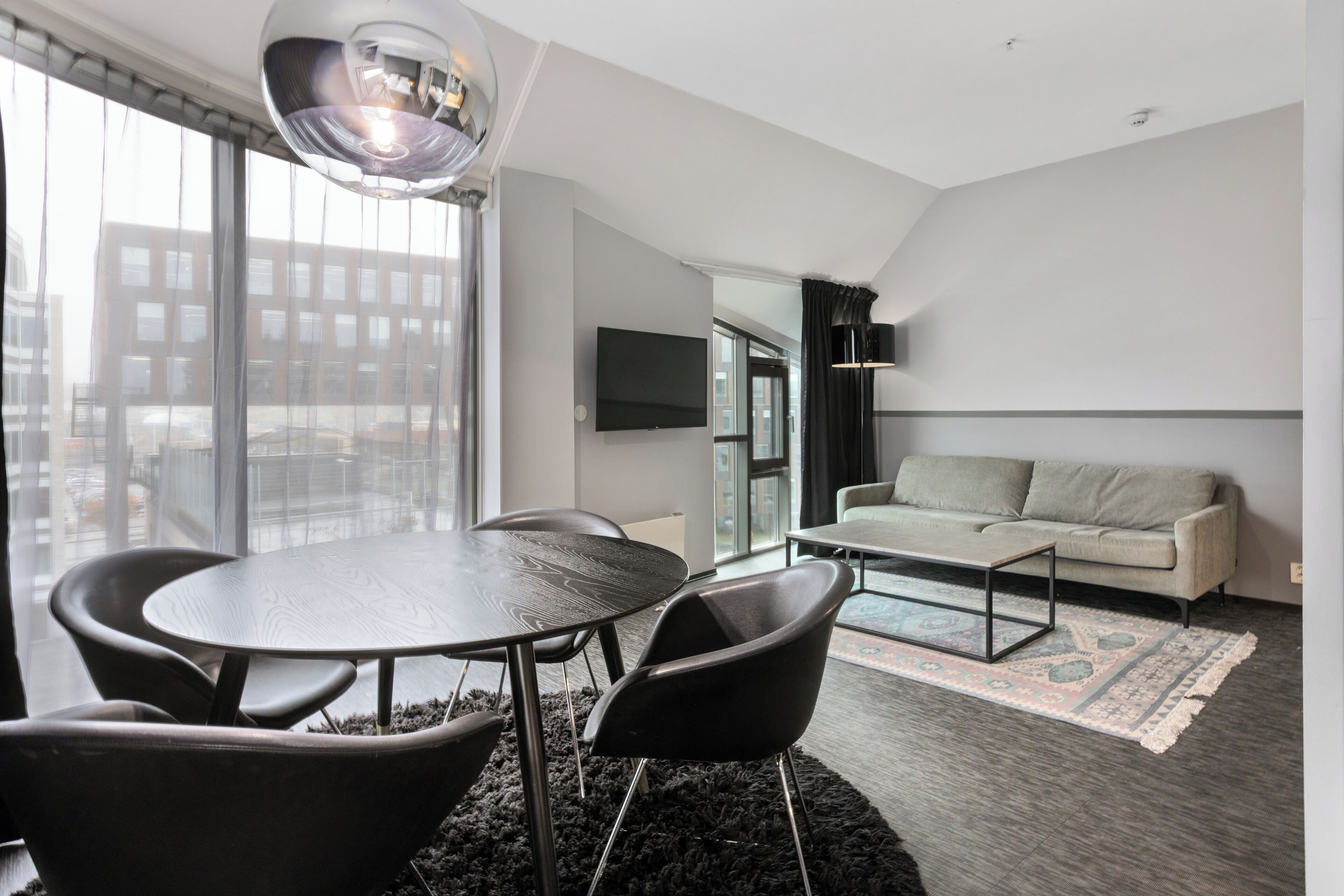 A modern living room with a round table, black chairs, a sofa, a TV mounted on the wall, and a large window with blinds.