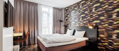 A modern hotel room with a large bed, a desk with a chair, and a view of the city through the window.