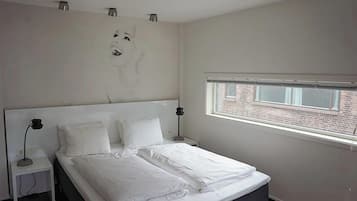 A bedroom with a large bed, two bedside tables, a window with a view of a brick building, and a wall-mounted light fixture.