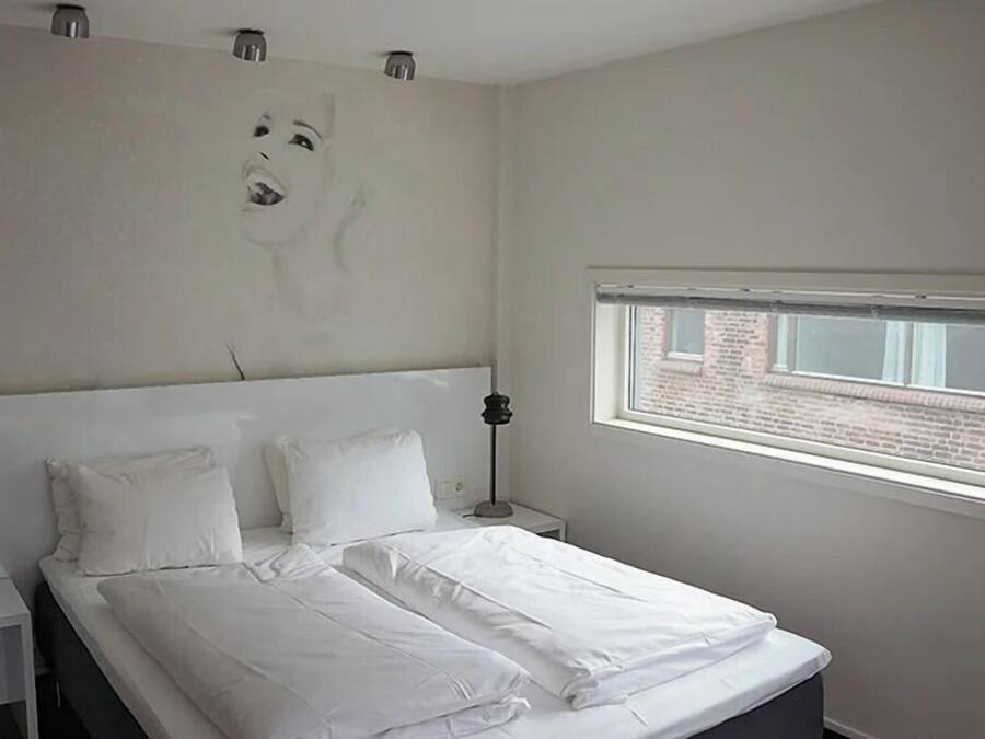 A bedroom with a large bed, two bedside tables, a window with a view of a brick building, and a wall-mounted light fixture.