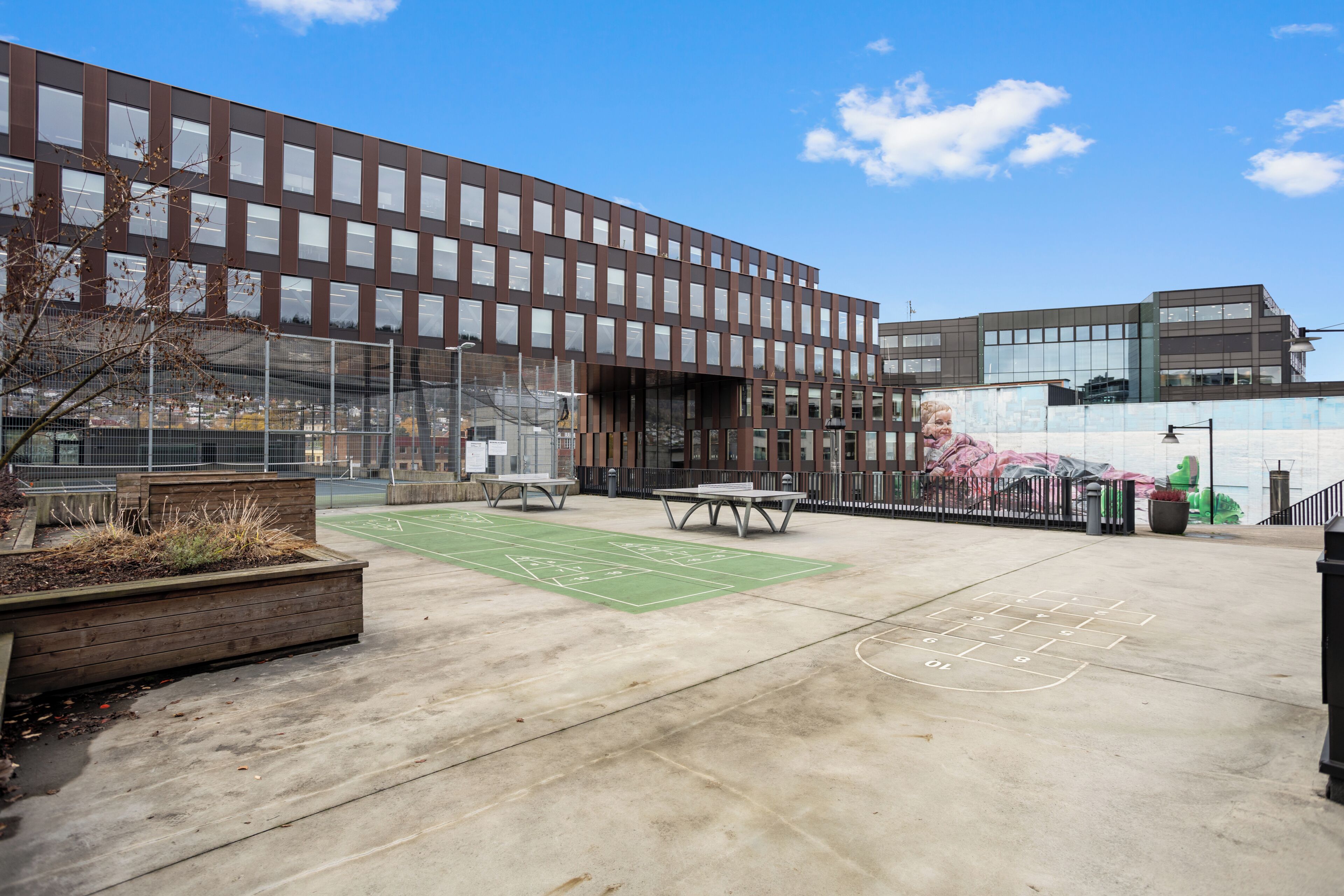 Un edificio moderno con un patio que incluye un tablero de rayuela, zonas de estar y un campo deportivo.