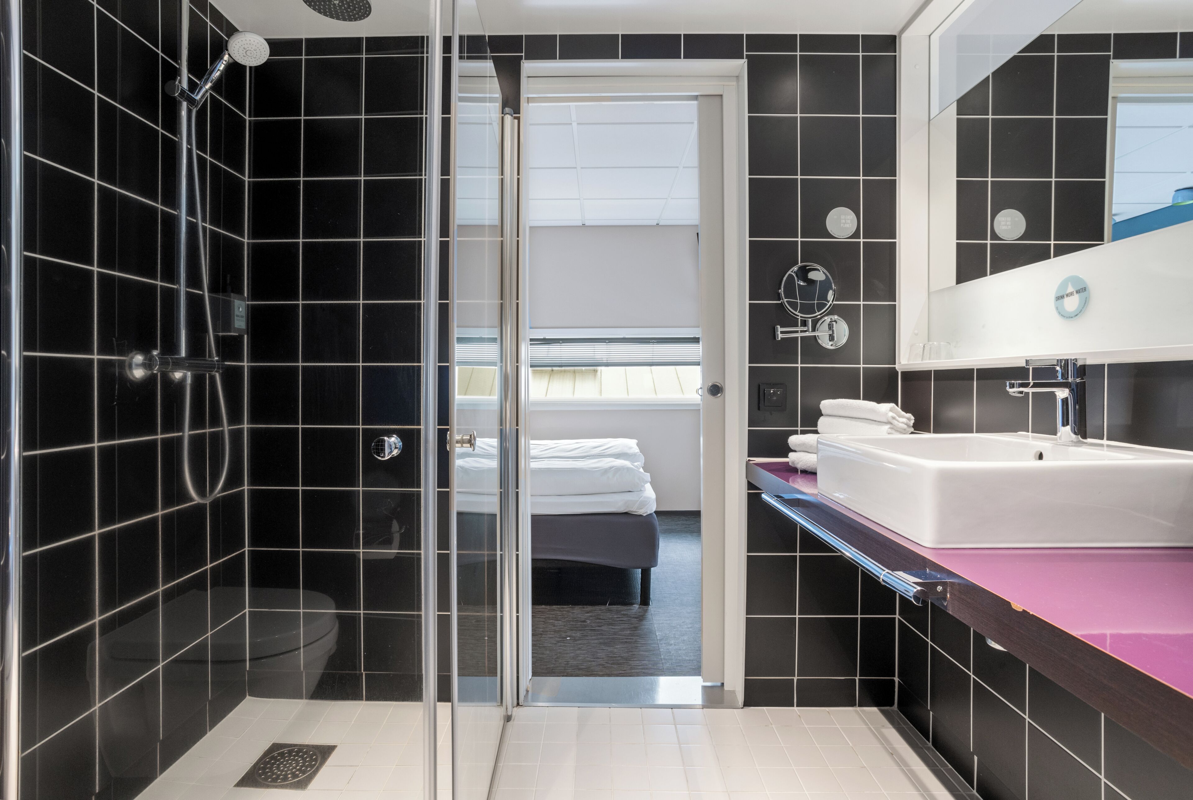 A modern bathroom with a glass-enclosed shower, a white sink, and a visible bedroom with a bed and window.