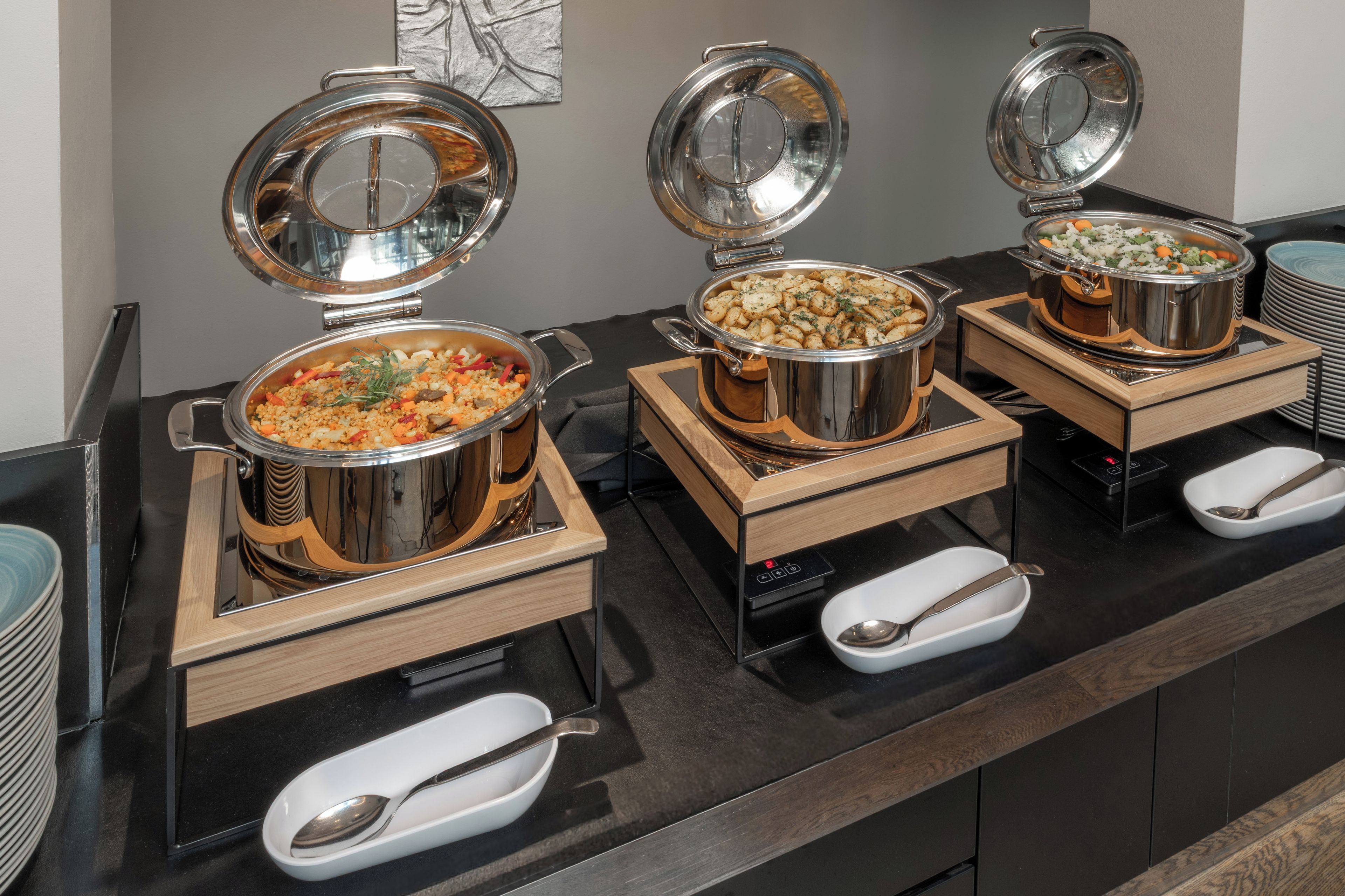 A buffet setup with multiple stainless steel chafing dishes on wooden stands, each with a lid and a spoon.